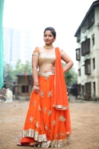A woman is standing confidently outdoors, wearing a traditional Indian lehenga choli. The outfit is bright orange with intricate gold embroidery and detailing. She accessorizes with gold jewelry, including a choker necklace and bangles. The background features an urban setting with buildings and greenery.