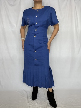 A person wearing a blue dress with button details and short sleeves, featuring pleats at the bottom. The dress is complemented by black ankle boots and a neutral wrist accessory.