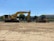 A yellow excavator and a yellow bulldozer are parked on a dirt construction site. The excavator bears the Komatsu brand and has 'PC490' printed on it. There is a building with a grey facade and trees in the background, as well as clear skies and some overhead utility lines.