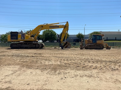 A yellow excavator and a yellow bulldozer are parked on a dirt construction site. The excavator bears the Komatsu brand and has 'PC490' printed on it. There is a building with a grey facade and trees in the background, as well as clear skies and some overhead utility lines.
