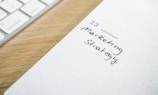 A close-up of a desk scene featuring part of a keyboard on the left and an open planner or diary on the right. The planner shows the date as Wednesday the 23rd, with handwritten text below it saying 'Marketing Strategy'. The scene is on a light wooden surface, creating a professional and organized atmosphere.