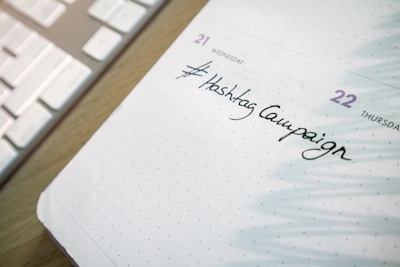 A close-up of a planner or calendar page showing handwritten text reading '#Hashtag Campaign'. The background includes a keyboard, suggesting a workspace setting.