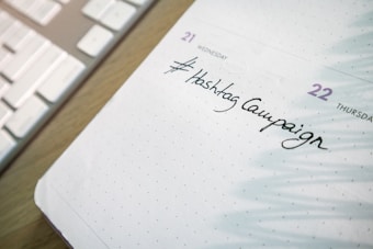 A close-up of a planner or calendar page showing handwritten text reading '#Hashtag Campaign'. The background includes a keyboard, suggesting a workspace setting.