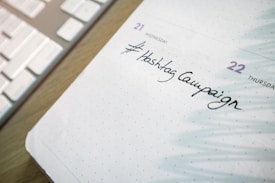 A close-up of a planner or calendar page showing handwritten text reading '#Hashtag Campaign'. The background includes a keyboard, suggesting a workspace setting.