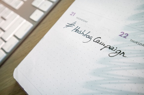 A close-up of a planner or calendar page showing handwritten text reading '#Hashtag Campaign'. The background includes a keyboard, suggesting a workspace setting.