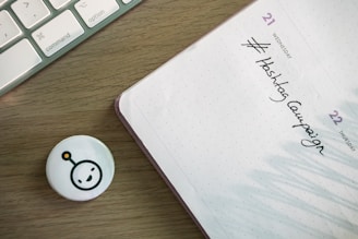 A wooden desk features a corner of a keyboard on the left and a planner on the right. The planner is opened to a page showing Wednesday the 21st and Thursday the 22nd with 'Hashtag Campaign' handwritten under Wednesday. A small circular badge with a smiling face icon lies near the planner.
