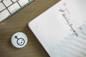 A wooden desk features a corner of a keyboard on the left and a planner on the right. The planner is opened to a page showing Wednesday the 21st and Thursday the 22nd with 'Hashtag Campaign' handwritten under Wednesday. A small circular badge with a smiling face icon lies near the planner.
