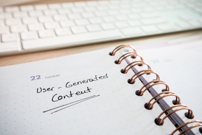 A close-up of a spiral-bound notebook open to a page with 'User-Generated Content' written on it in black ink. The page indicates it's Tuesday, the 22nd, and the notebook lies on a desk beside a white computer keyboard.