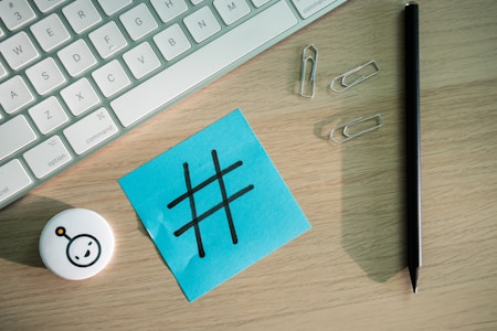 A computer keyboard is partially visible alongside a blue sticky note featuring a large black hashtag symbol. Nearby, several silver paperclips, a black pen, and a small round object with a simple face icon on it are present. The surface appears to be a light wood desk.