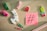 A variety of colorful stationery items and small toys are arranged on a light wooden surface, including a white llama with a colorful design, vibrant clothespins, paperclips, a red geometric shape, and a cactus. A pink sticky note with a black hashtag symbol is also present.