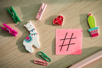 A variety of colorful stationery items and small toys are arranged on a light wooden surface, including a white llama with a colorful design, vibrant clothespins, paperclips, a red geometric shape, and a cactus. A pink sticky note with a black hashtag symbol is also present.