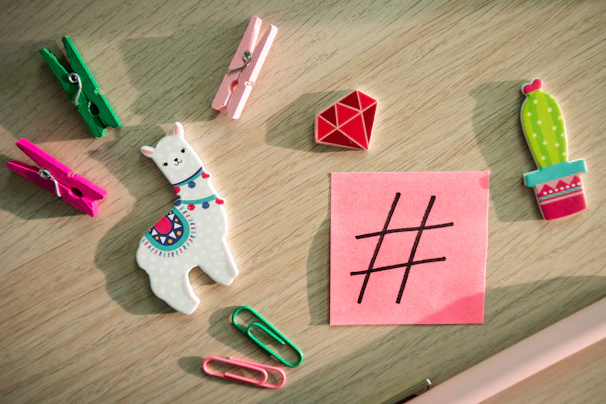 A variety of colorful stationery items and small toys are arranged on a light wooden surface, including a white llama with a colorful design, vibrant clothespins, paperclips, a red geometric shape, and a cactus. A pink sticky note with a black hashtag symbol is also present.