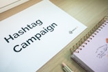 A sheet of paper with the words 'Hashtag Campaign' printed on it is laid on a wooden surface. Next to it is a spiral-bound notebook with a drawing on the cover, and a pencil. The logo 'WALLS.IO' is visible on the paper.