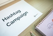 A sheet of paper with the words 'Hashtag Campaign' printed on it is laid on a wooden surface. Next to it is a spiral-bound notebook with a drawing on the cover, and a pencil. The logo 'WALLS.IO' is visible on the paper.