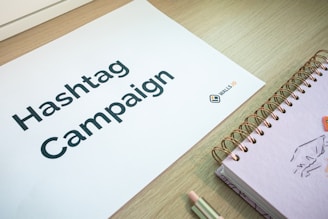 A sheet of paper with the words 'Hashtag Campaign' printed on it is laid on a wooden surface. Next to it is a spiral-bound notebook with a drawing on the cover, and a pencil. The logo 'WALLS.IO' is visible on the paper.