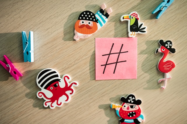 A collection of colorful cartoon-themed magnets and clothespins arranged on a wooden surface around a sticky note with a large hashtag symbol. The magnets include pirate-themed animals and characters like an octopus and birds wearing pirate hats.
