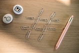 A collection of office supplies on a wooden surface, including several paperclips arranged in a tic-tac-toe grid pattern, two round pins with abstract designs, and a pink pen with gold accents.