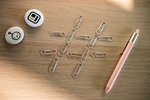 A collection of office supplies on a wooden surface, including several paperclips arranged in a tic-tac-toe grid pattern, two round pins with abstract designs, and a pink pen with gold accents.