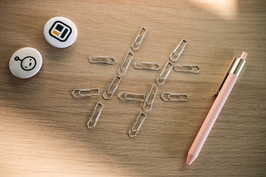 A collection of office supplies on a wooden surface, including several paperclips arranged in a tic-tac-toe grid pattern, two round pins with abstract designs, and a pink pen with gold accents.