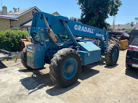 A large blue telescopic forklift labeled 'GRADALL 534D-9' with oversized tires is parked outdoors on a gravel surface. Nearby, there is a black vehicle partially visible, along with greenery and residential buildings in the background. The area is well-lit by sunlight and the surrounding environment has various construction materials.