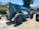 A large blue telescopic forklift labeled 'GRADALL 534D-9' with oversized tires is parked outdoors on a gravel surface. Nearby, there is a black vehicle partially visible, along with greenery and residential buildings in the background. The area is well-lit by sunlight and the surrounding environment has various construction materials.