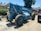 A large blue telescopic forklift labeled 'GRADALL 534D-9' with oversized tires is parked outdoors on a gravel surface. Nearby, there is a black vehicle partially visible, along with greenery and residential buildings in the background. The area is well-lit by sunlight and the surrounding environment has various construction materials.