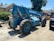 A large blue telescopic forklift labeled 'GRADALL 534D-9' with oversized tires is parked outdoors on a gravel surface. Nearby, there is a black vehicle partially visible, along with greenery and residential buildings in the background. The area is well-lit by sunlight and the surrounding environment has various construction materials.