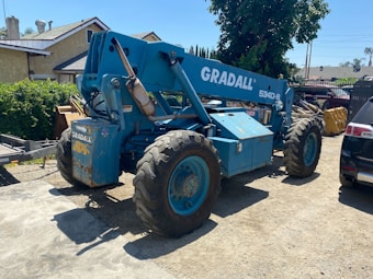 A large blue telescopic forklift labeled 'GRADALL 534D-9' with oversized tires is parked outdoors on a gravel surface. Nearby, there is a black vehicle partially visible, along with greenery and residential buildings in the background. The area is well-lit by sunlight and the surrounding environment has various construction materials.