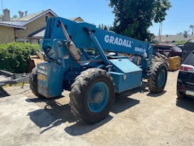 A large blue telescopic forklift labeled 'GRADALL 534D-9' with oversized tires is parked outdoors on a gravel surface. Nearby, there is a black vehicle partially visible, along with greenery and residential buildings in the background. The area is well-lit by sunlight and the surrounding environment has various construction materials.