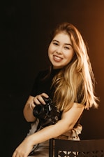 A warm portrait of Tatiane Morais holding a camera, smiling gently in a cozy studio setting.