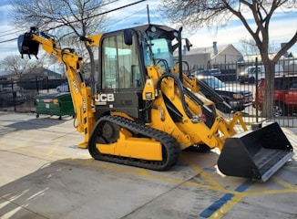 A yellow excavator with a large bucket attachment is parked on a concrete surface in an outdoor area. It has caterpillar tracks for movement and hydraulic pipes running along its arm. There are bare trees and a fenced area with parked vehicles in the background.
