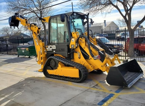 A yellow excavator with a large bucket attachment is parked on a concrete surface in an outdoor area. It has caterpillar tracks for movement and hydraulic pipes running along its arm. There are bare trees and a fenced area with parked vehicles in the background.