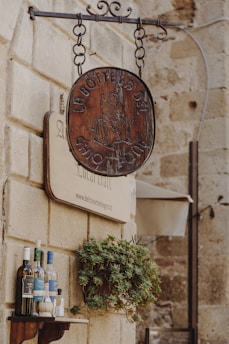 A rustic wooden sign hangs from a metal bracket against a stone wall. Below it, several wine bottles are neatly arranged on a small wooden shelf beside a potted plant. The stone wall gives a historic and quaint atmosphere.