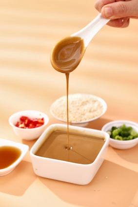 A hand is holding a white spoon pouring a thick, brown sauce into a square white dish filled with the same sauce. Surrounding the dish are small bowls containing minced red chili, chopped green onions, and a pile of sesame seeds. The background features a light orange surface with soft shadows.