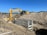 Two construction workers wearing helmets and high-visibility vests are on a construction site with sandy terrain. An excavator is actively digging a trench, and large mounds of earth surround the machinery. The sky is clear with no visible clouds.