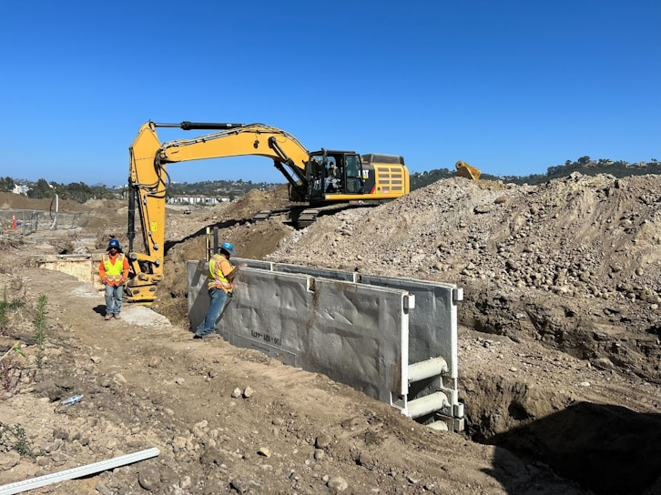 Two construction workers wearing helmets and high-visibility vests are on a construction site with sandy terrain. An excavator is actively digging a trench, and large mounds of earth surround the machinery. The sky is clear with no visible clouds.