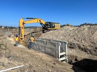 Two construction workers wearing helmets and high-visibility vests are on a construction site with sandy terrain. An excavator is actively digging a trench, and large mounds of earth surround the machinery. The sky is clear with no visible clouds.