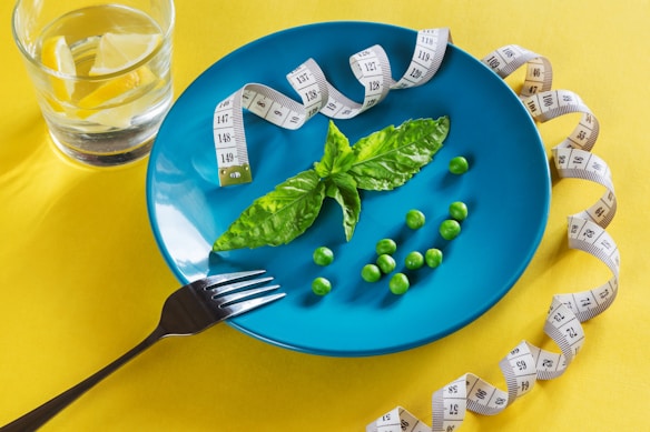 A vibrant blue plate contains a few green peas and a couple of basil leaves. A measuring tape is loosely arranged around the plate, and a glass of water with lemon slices sits nearby. A silver fork rests on the edge of the plate.