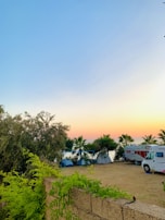 A serene outdoor camping scene at sunset with a clear blue sky transitioning to orange at the horizon. Several tents are set up among palm trees near a RV, and lush green foliage and trees surround the area.