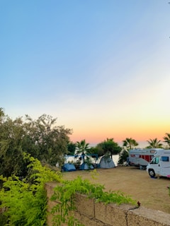 A serene outdoor camping scene at sunset with a clear blue sky transitioning to orange at the horizon. Several tents are set up among palm trees near a RV, and lush green foliage and trees surround the area.