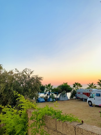 A serene outdoor camping scene at sunset with a clear blue sky transitioning to orange at the horizon. Several tents are set up among palm trees near a RV, and lush green foliage and trees surround the area.