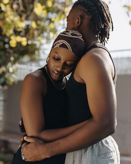 A couple is seen embracing each other tenderly. The woman, with closed eyes and a peaceful expression, wears a headscarf and has a nose ring, while the man has his back towards the camera, showing his dreadlocks. Sunlight filters through the background tree, casting a gentle light on the scene.