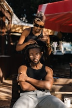 A woman with sunglasses is styling a man's hair outdoors under a red and white canopy. The man, seated on a wooden bench, has his arms crossed and is looking directly at the camera.