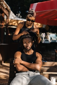 A woman with sunglasses is styling a man's hair outdoors under a red and white canopy. The man, seated on a wooden bench, has his arms crossed and is looking directly at the camera.