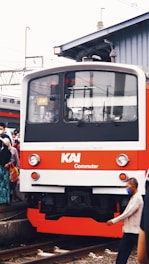 A commuter train with a red and white exterior is at a rail platform, surrounded by people. Some individuals are wearing face masks. The scene is set in an urban area with overhead wires and a building in the background.
