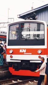 A commuter train with a red and white exterior is at a rail platform, surrounded by people. Some individuals are wearing face masks. The scene is set in an urban area with overhead wires and a building in the background.