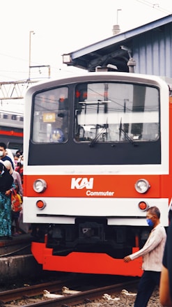A commuter train with a red and white exterior is at a rail platform, surrounded by people. Some individuals are wearing face masks. The scene is set in an urban area with overhead wires and a building in the background.