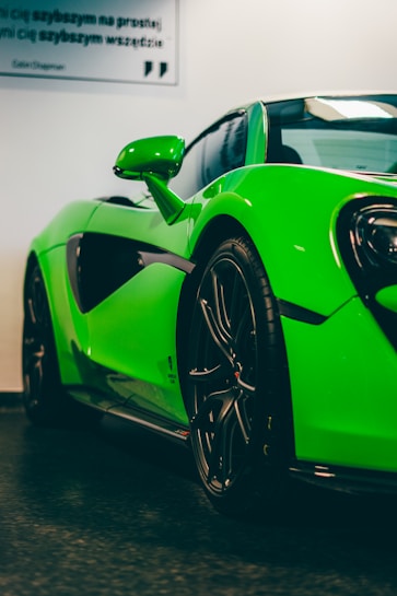 A vibrant green sports car, viewed at an angle from the front side. The car is sleek and modern with black alloy wheels. It is parked indoors with a smooth floor, and part of the background shows a white wall with a quote in Polish.