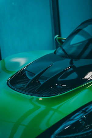 A close-up of a car's hood with sleek design lines and visible vents. The paint is a bright, lime green with reflective surfaces giving it a glossy appearance. The background is an out-of-focus, cool-toned interior space, adding contrast to the vividness of the car.