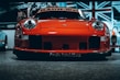 A vibrant red sports car with a sleek and aggressive design is pictured from the front, prominently displaying its headlights and aerodynamic features. The background includes a large Union Jack flag and additional vehicles, indicating an indoor exhibition or showroom setting.